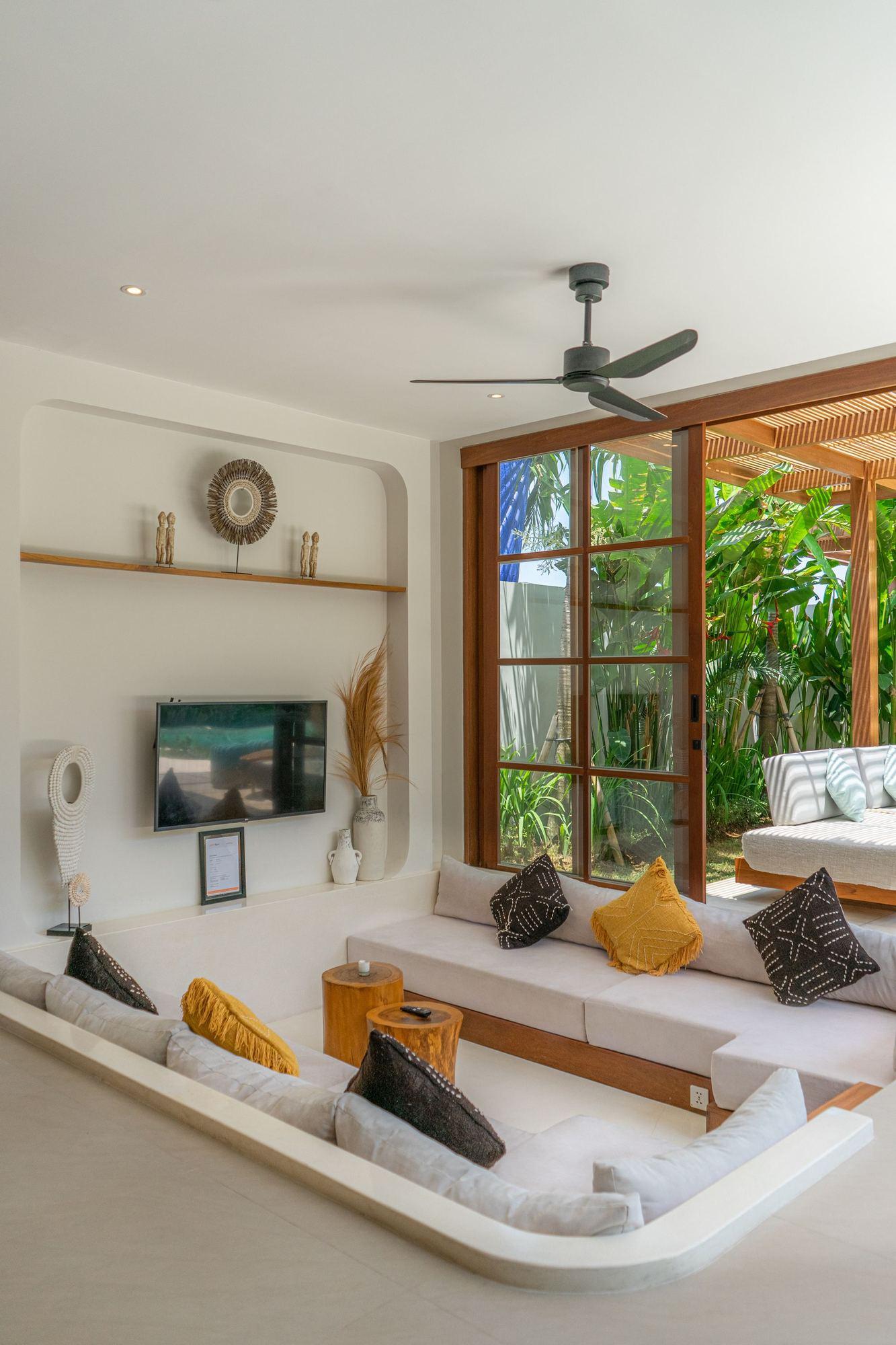 A bright living room with a ceiling fan, cushioned seating, and wood-framed glass doors open to greenery.