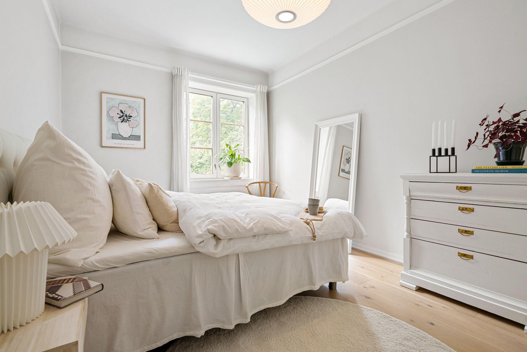 A bright bedroom with white bedding, soft pillows, and daylight through a tall window.